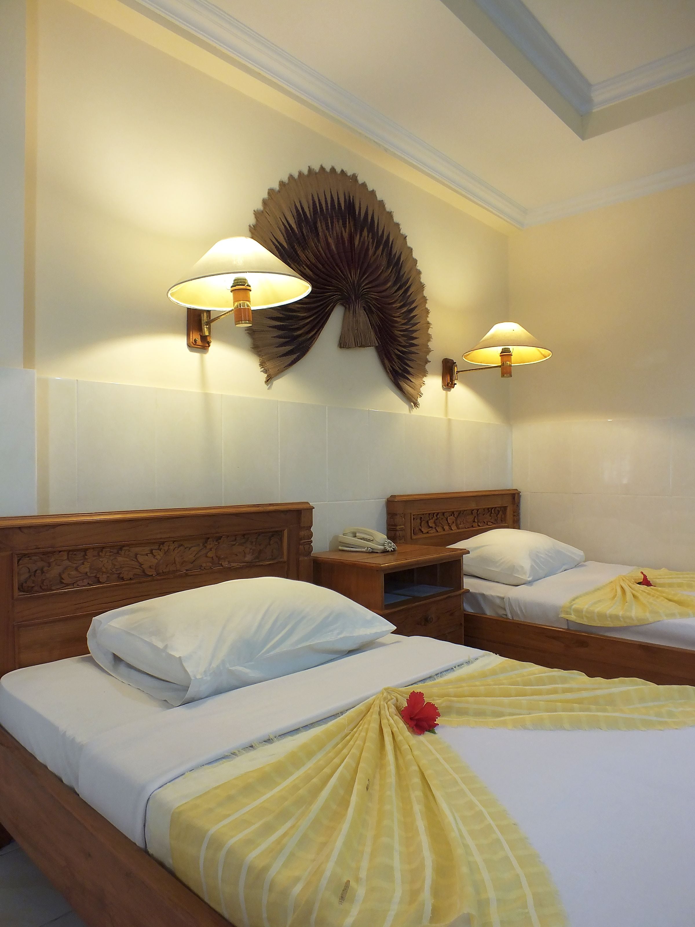 A hotel room with two wooden beds, white linens, a decorative wall piece, and wall-mounted lamps.