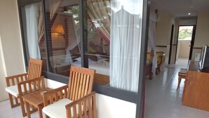 Una habitación con un ventanal grande, sillas de madera, una mesita y una cama con colcha blanca.