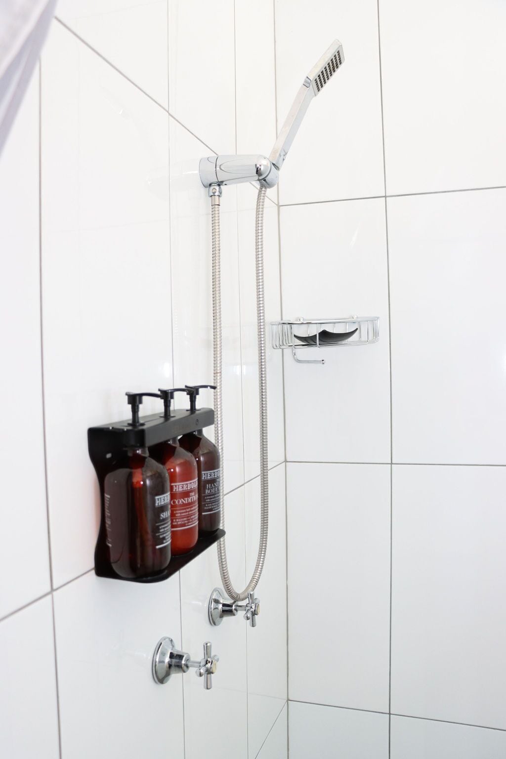 Ducha con cabezal de ducha de mano y extensión de ducha manual. Hay tres botellas de champú y acondicionador en un estante debajo de la ducha.