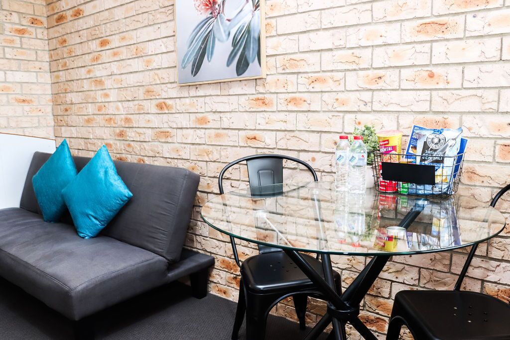 A modern room with a brick wall, a glass table, a grey sofa with blue cushions, and a metal chair.