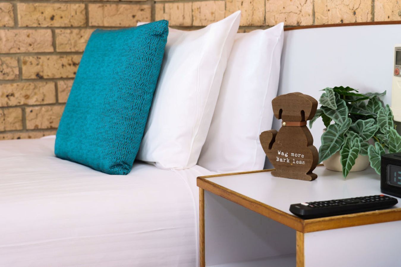 Un letto con cuscini bianchi e color acquamarina, una piccola statuina di un orso di legno, una pianta e un telecomando su un comodino.