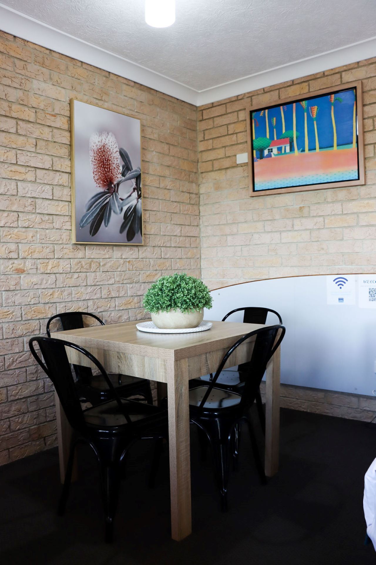 A dining area with a brick wall, a round wooden table, black chairs, a potted plant, and artwork.