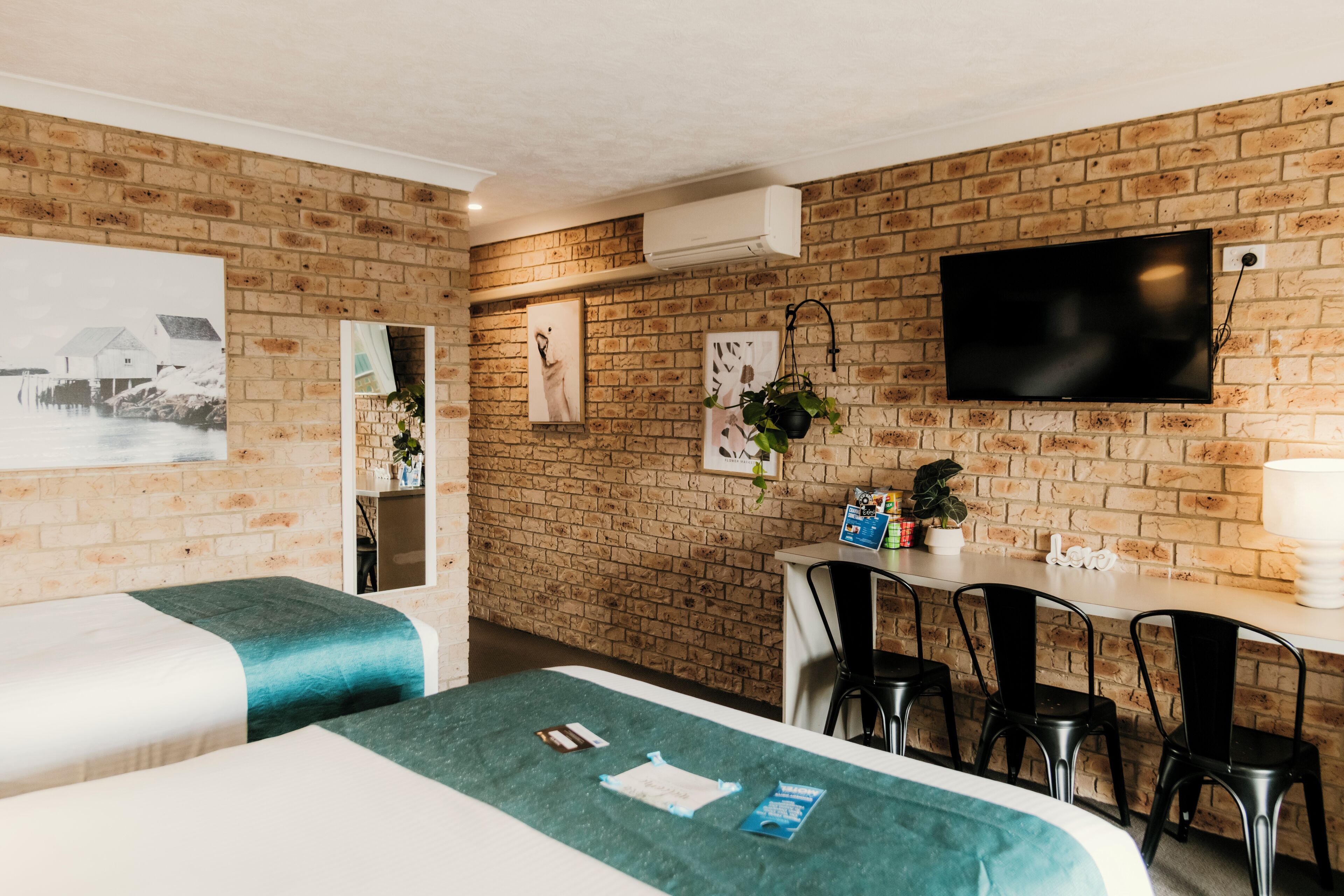 A hotel room with a brick wall, a flat-screen TV, two beds with teal bedding, a desk with a chair, and a small table with a plant.