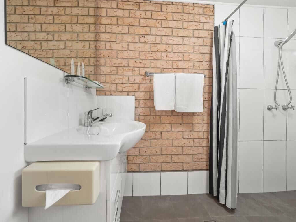 A bathroom with a brick wall, a white sink, a glass shelf, a towel rack, and a shower area.