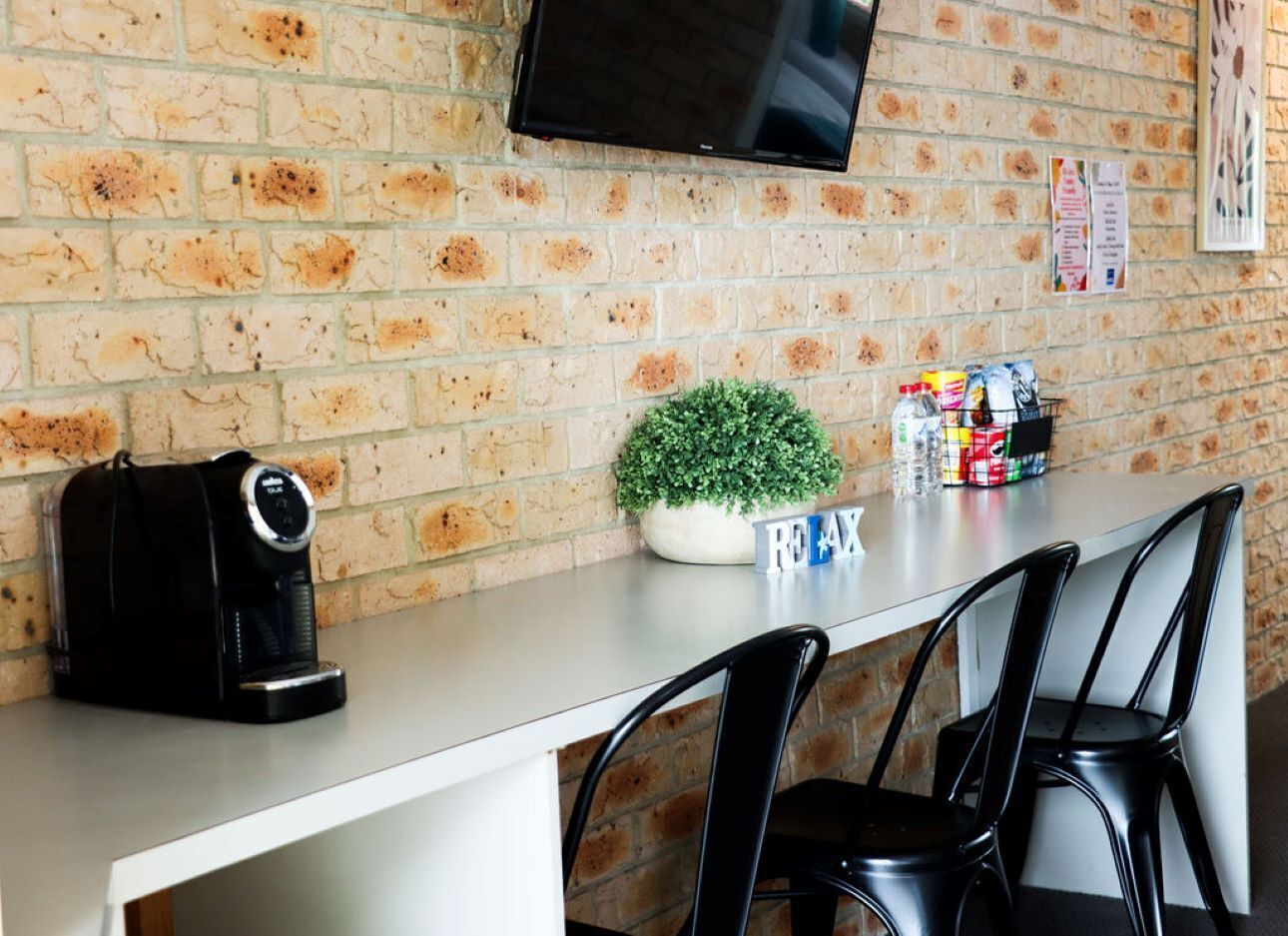 A modern bar area with a brick wall, a flat-screen TV mounted above a counter, a coffee machine, a small potted plant, and a selection of snacks and beverages.