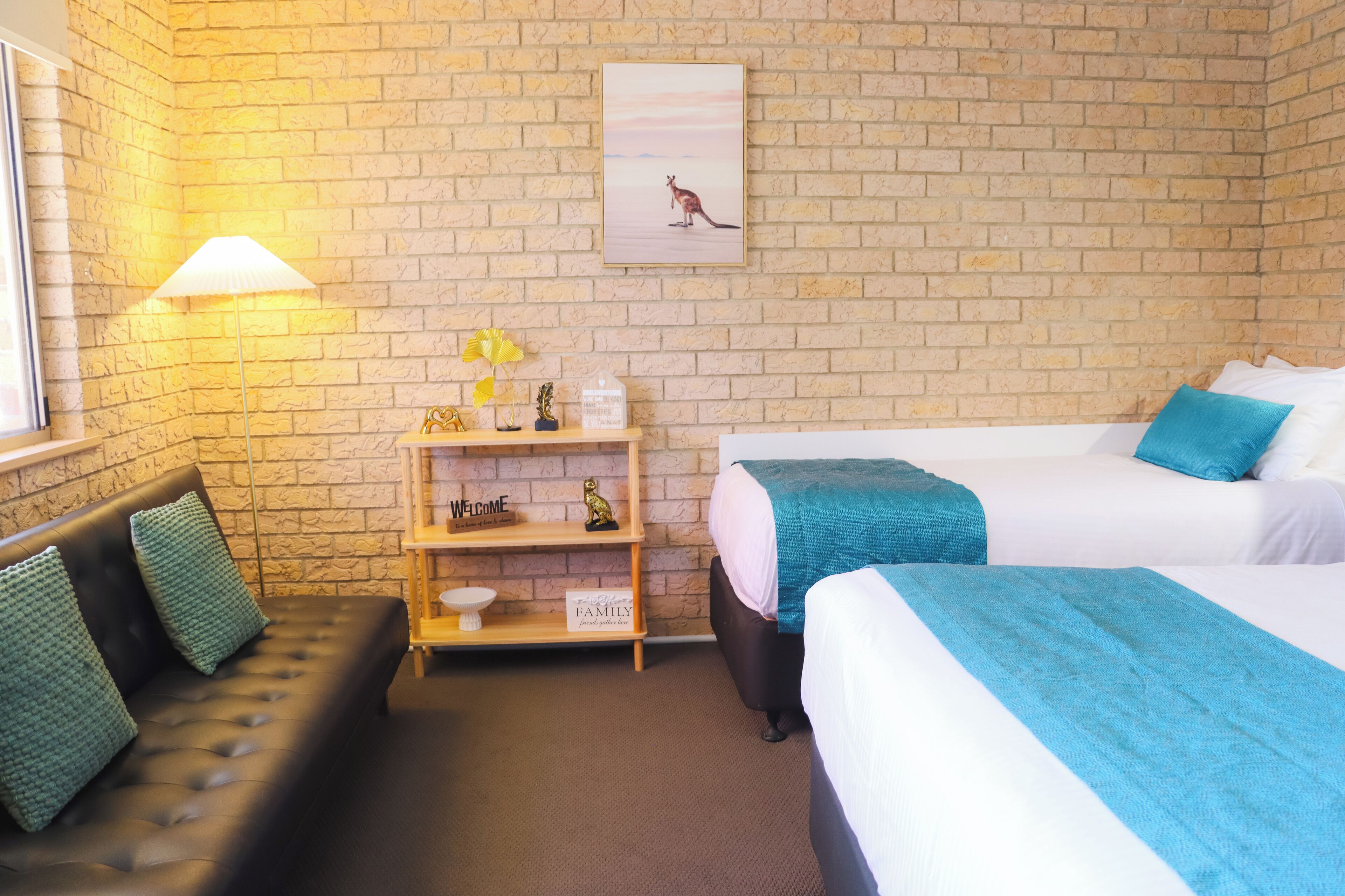 Une chambre d’hôtel avec un mur en briques, un canapé avec des coussins verts, deux lits avec une literie bleue et blanche, une table de chevet avec une lampe et un tableau encadré représentant un kangourou.