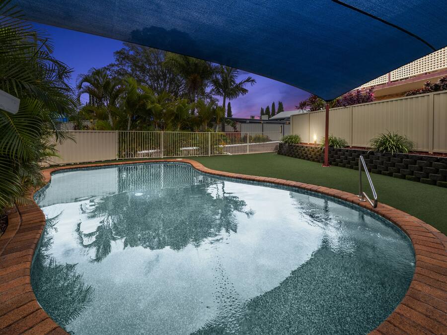 Una piscina con borde de ladrillo, rodeada de una valla y follaje.