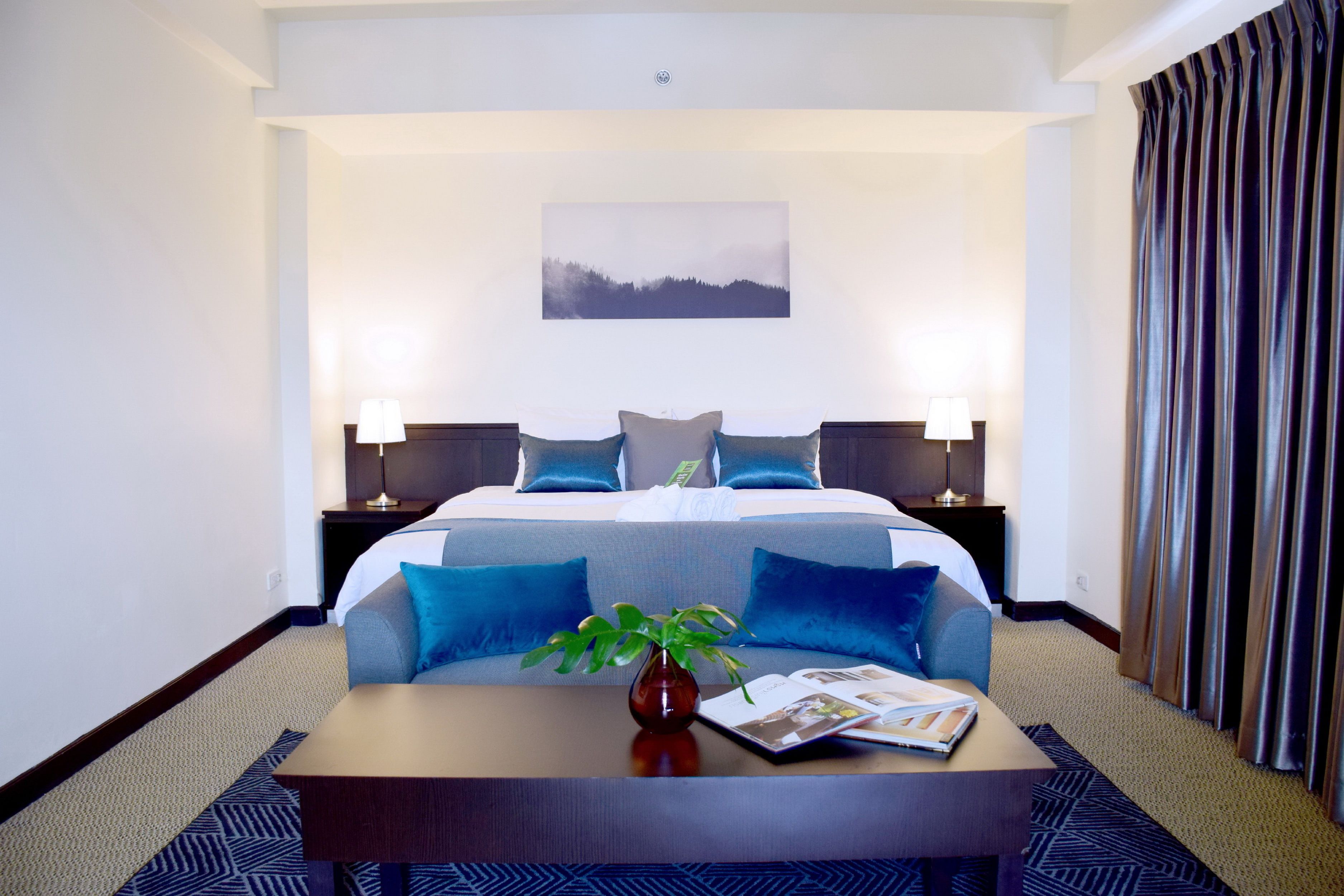 A hotel room with a large bed, two bedside tables with lamps, a coffee table with a plant and magazine, and a painting on the wall.