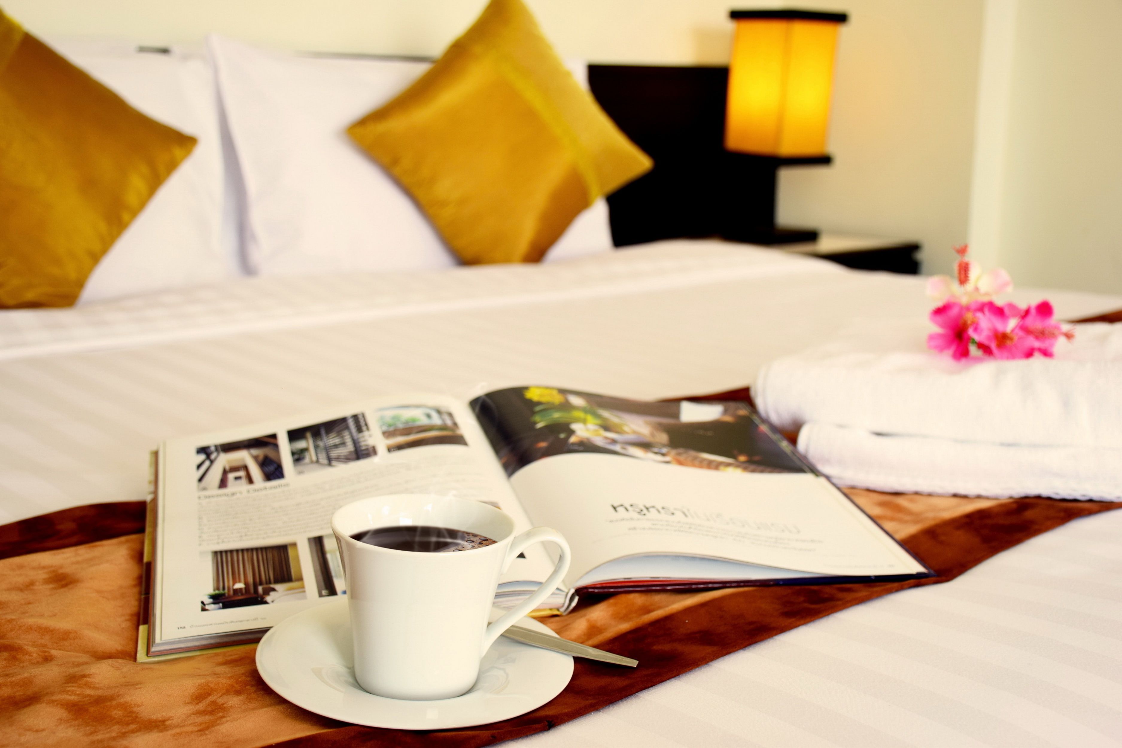 A neatly made bed with a white and gold pillow arrangement, a bedside lamp, a cup of coffee, a magazine, and a pink flower on a wooden tray.