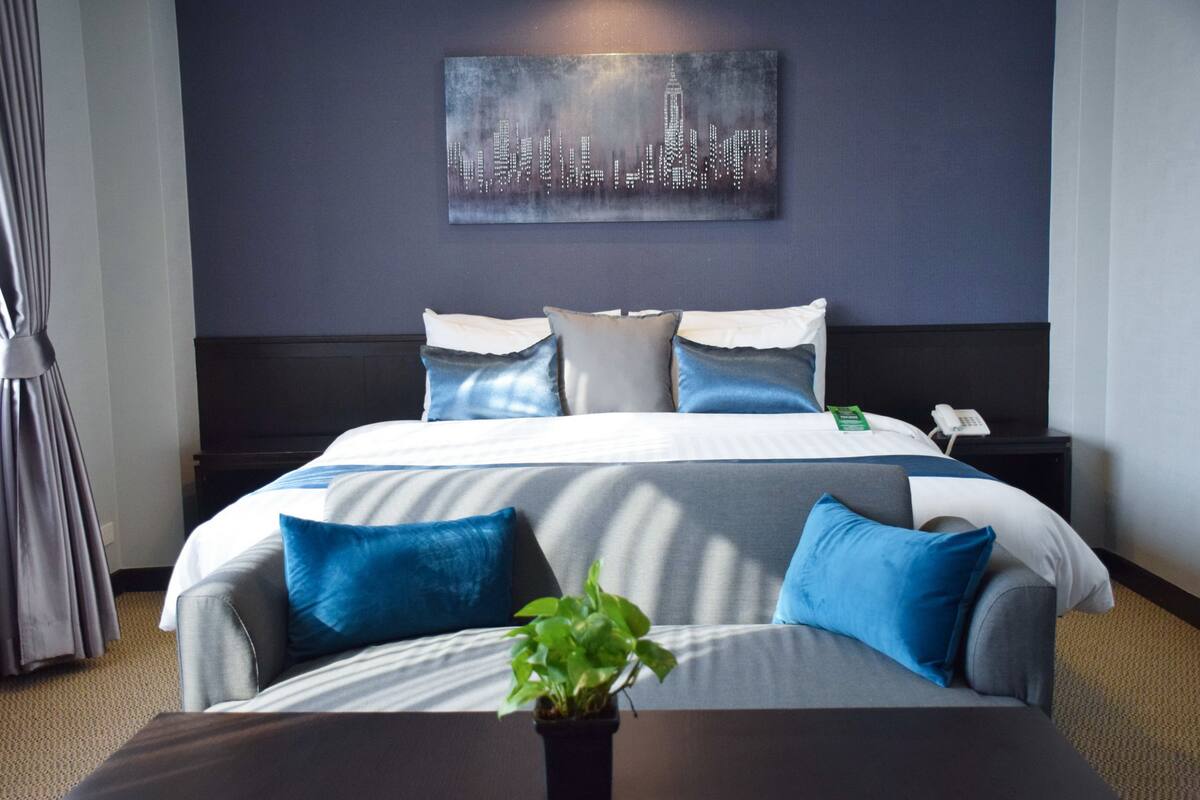 A hotel room with a bed, a nightstand, a plant, and a cityscape artwork on the wall.