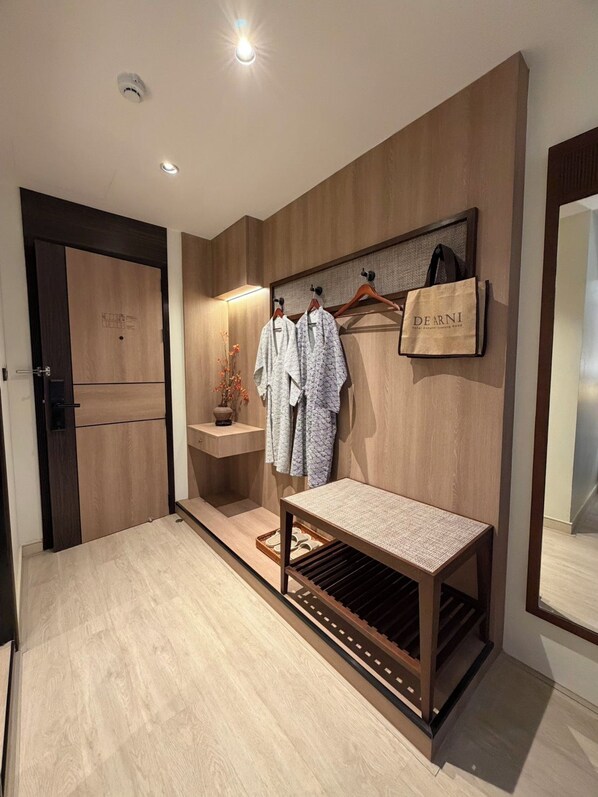 A hotel room with a wooden wardrobe, a bench, a towel rack with robes, and a small table with a vase of flowers. - De Arni Hotel Bangkok (Bangkok)