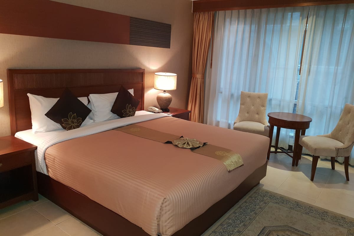 Een hotelkamer met een groot bed, een nachtkastje met een lamp, twee stoelen en uitzicht buiten door dunne gordijnen.