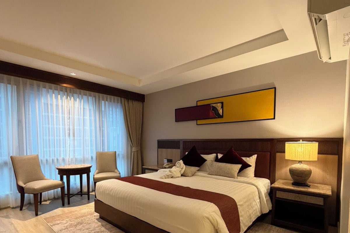 Habitación de hotel con una cama grande, una mesita de noche con lámpara, una silla y vistas al exterior a través de cortinas translúcidas.