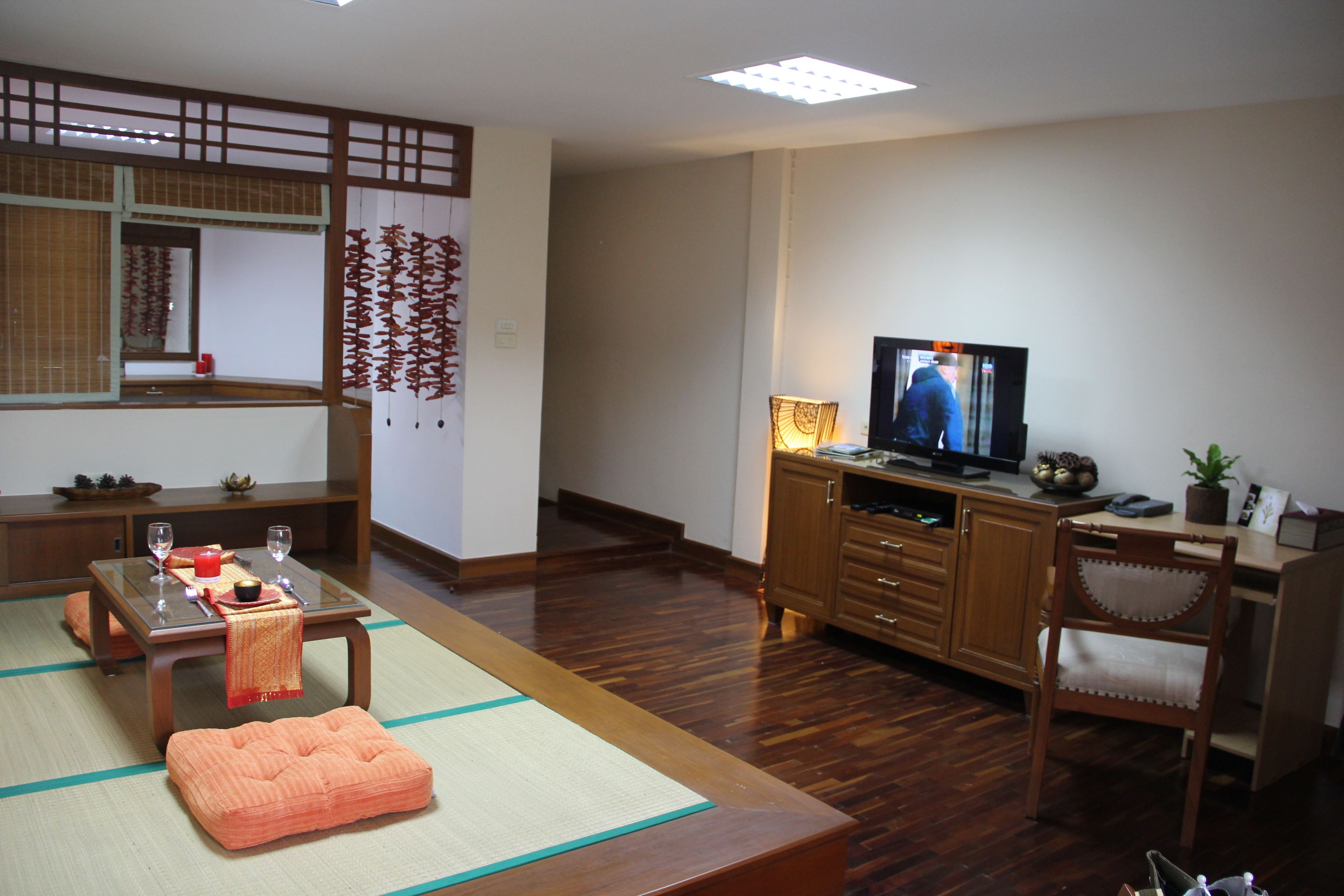 A traditional Japanese-style living room with tatami flooring, a low table with a red cloth, a television on a wooden cabinet, and a sliding door with decorative paper.