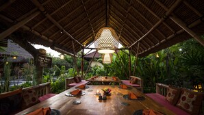 An outdoor dining area with a thatched roof, wooden tables, and cushioned seating. - Man's Cottages and Spa (Pemuteran)