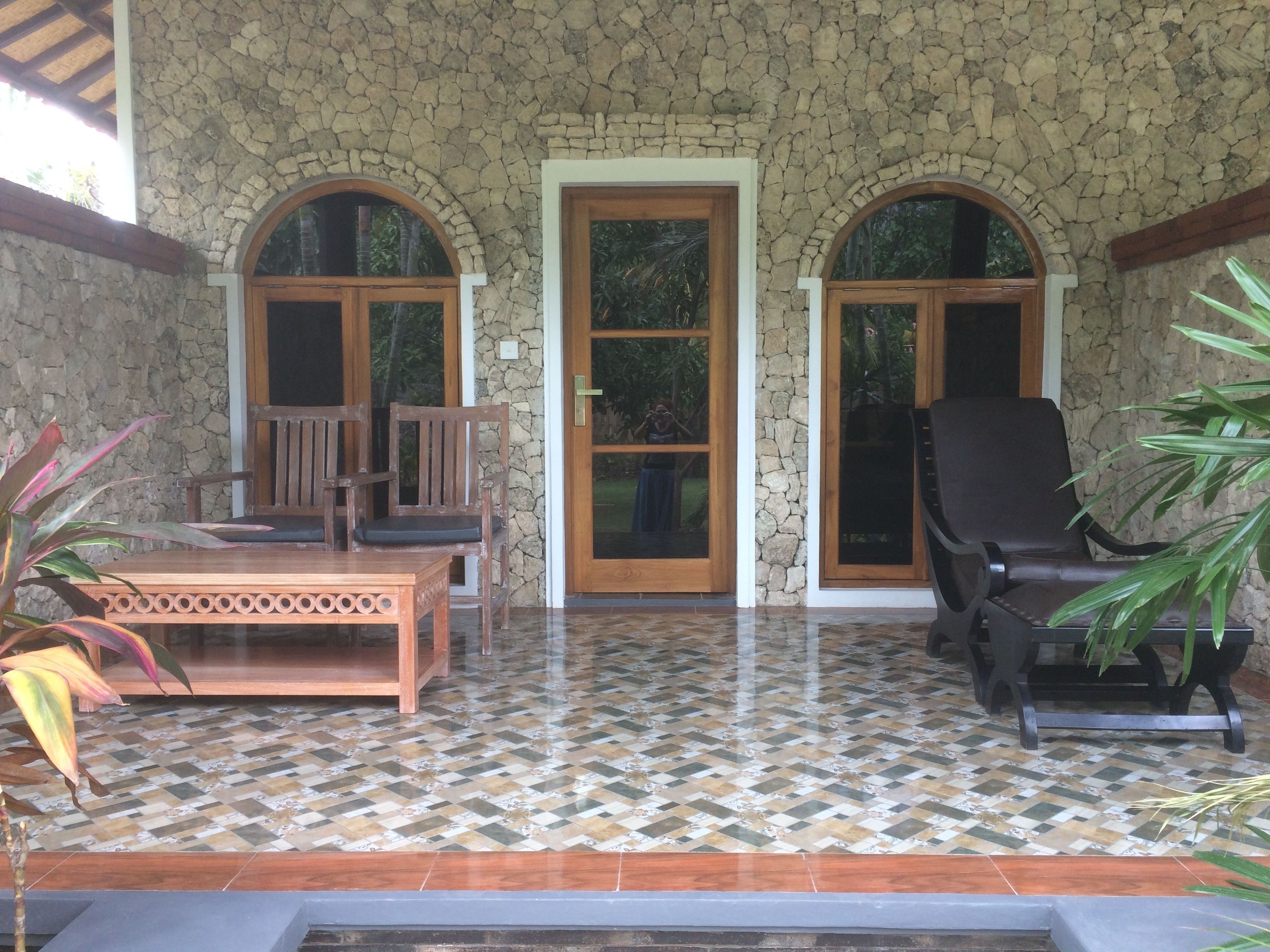 A covered patio with stone walls, a tiled floor, wooden furniture, and a stone door.