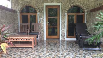 A covered patio with stone walls, a tiled floor, wooden furniture, and a stone door.