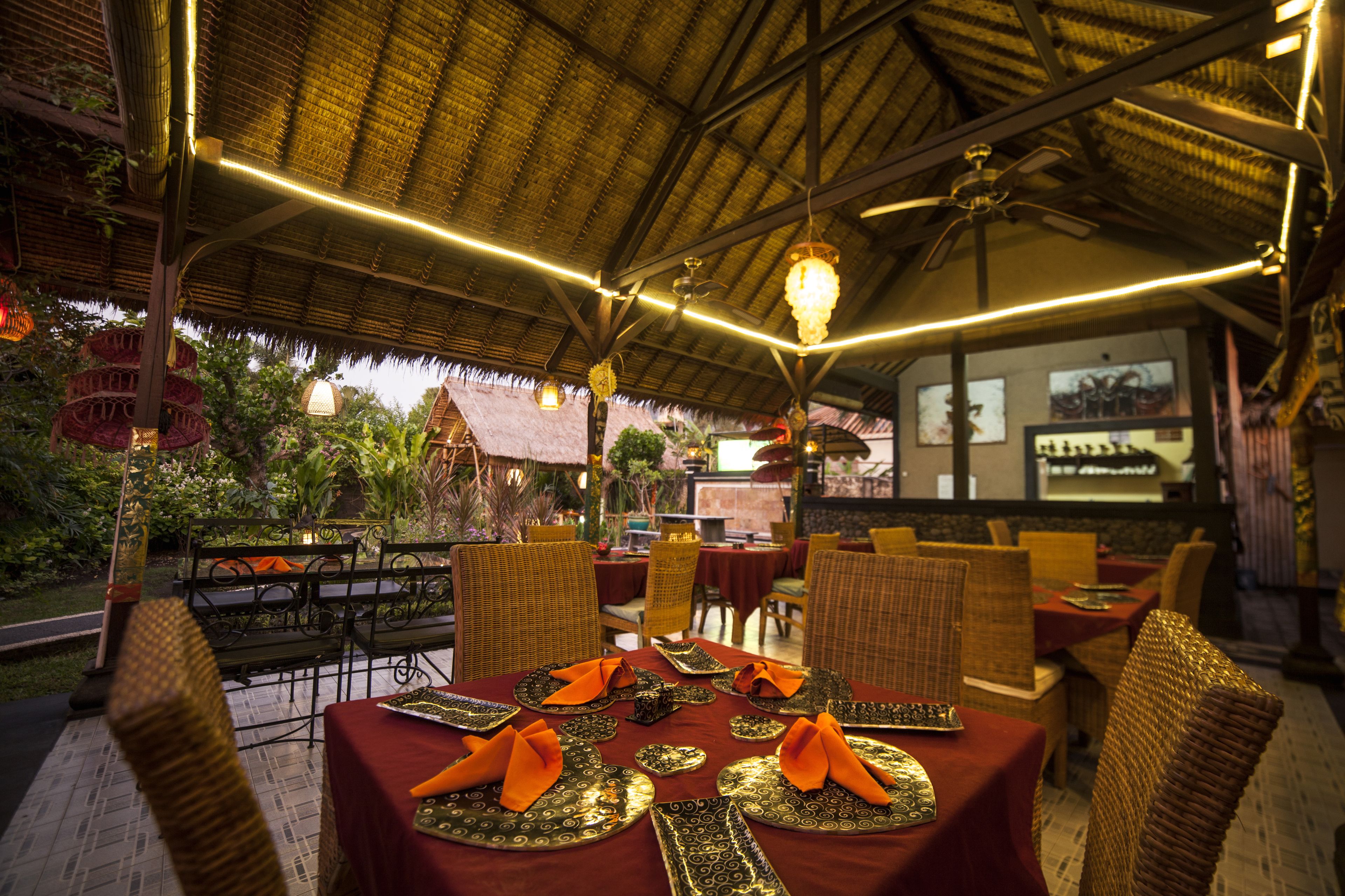 An outdoor restaurant with thatched roof, wicker furniture, and tables set with orange napkins and decorative plates.
