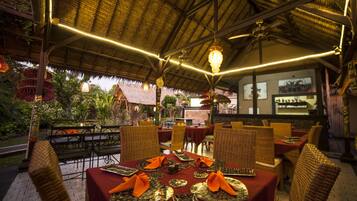 An outdoor restaurant with thatched roof, wicker furniture, and tables set with orange napkins and decorative plates.