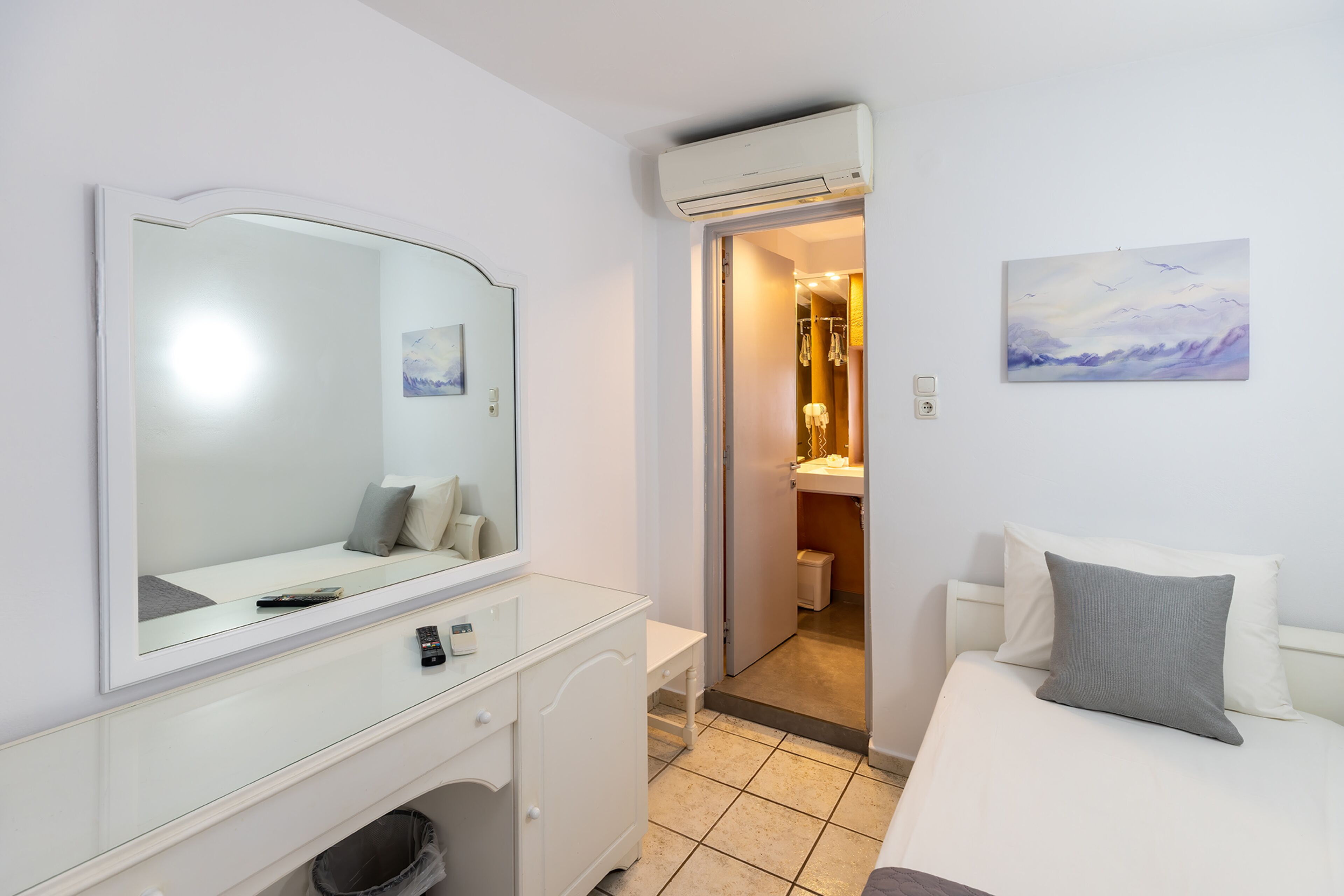 A hotel room with a large mirror, a bed with pillows, a bedside table, and a view of the bathroom.