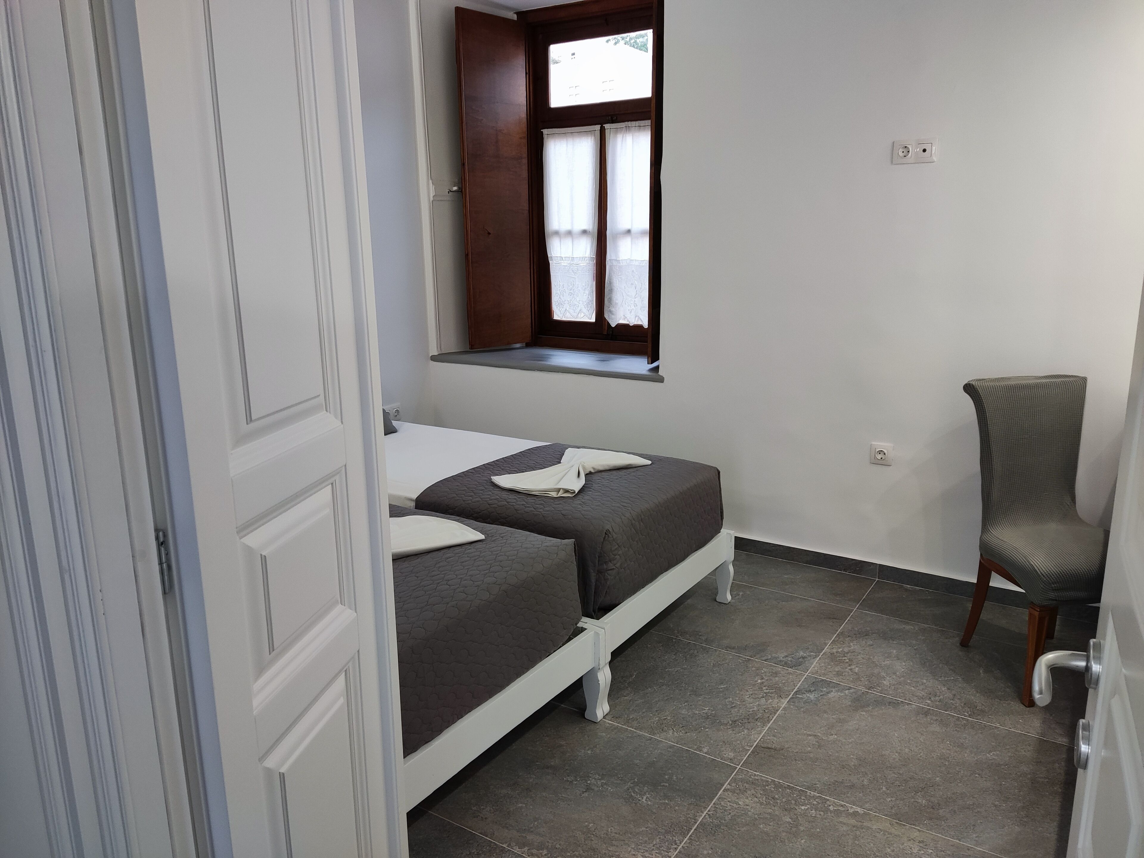 Una habitación pequeña y limpia con dos camas, una ventana con cortinas, una silla y piso de baldosas.