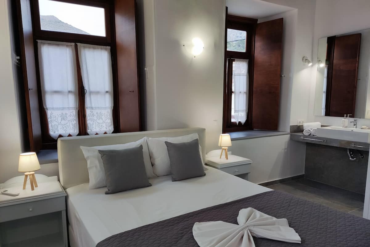 Une chambre d’hôtel avec un lit, des lampes de chevet, une salle de bain avec un lavabo et un miroir, et des fenêtres avec des rideaux.