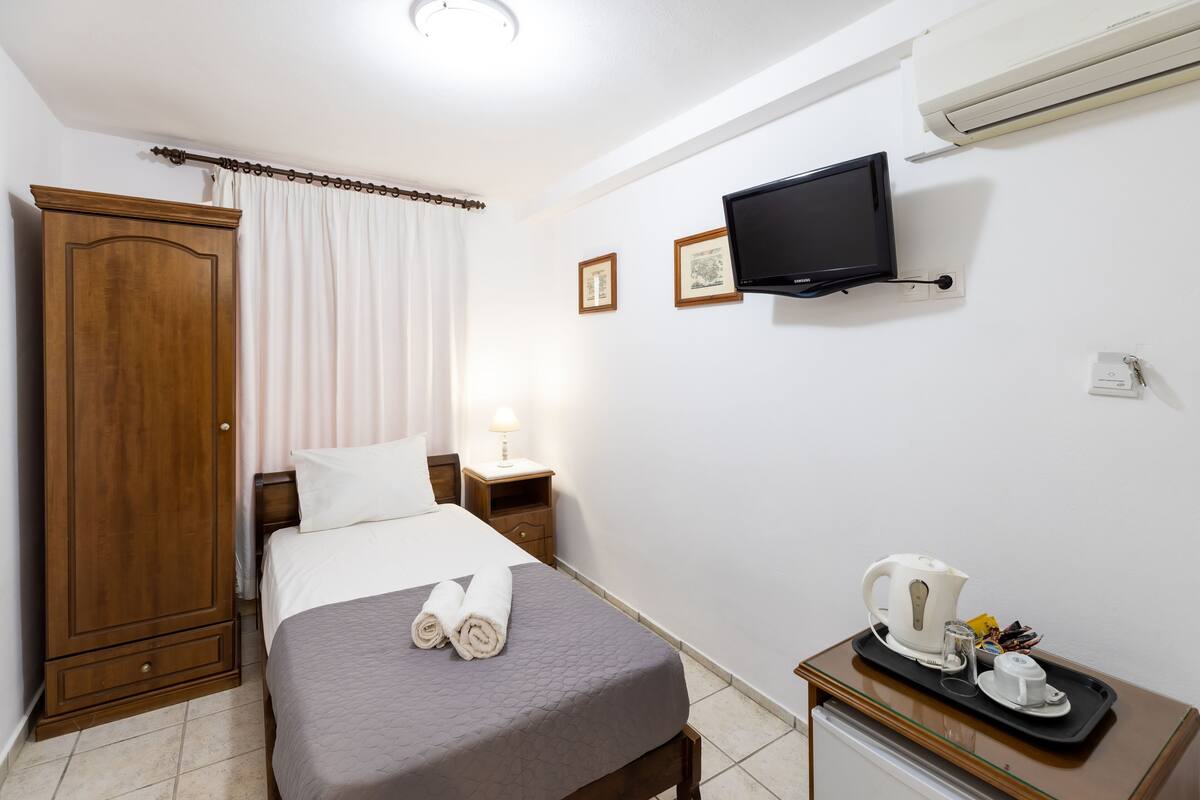 Une chambre d’hôtel comprenant un lit, une armoire en bois, une télévision, une table de chevet avec une lampe, et un plateau avec une bouilloire et des tasses.