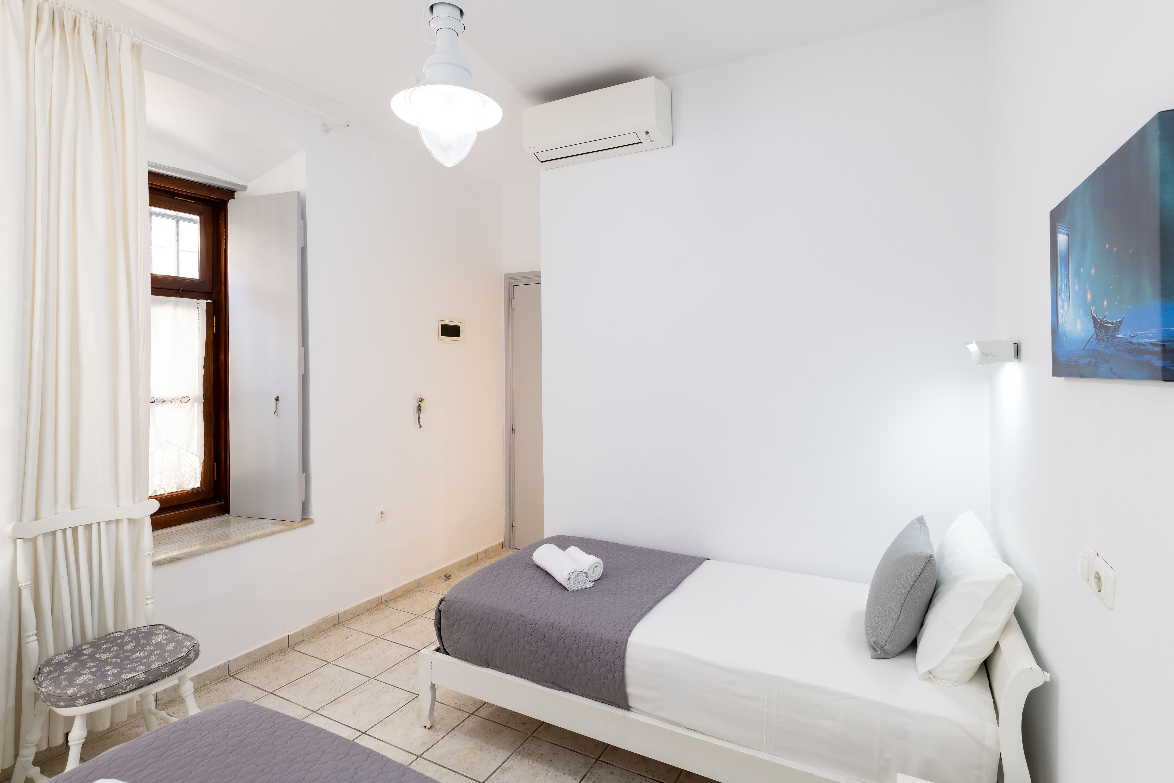 Un dormitorio con cama, una silla, una ventana con cortinas, un equipo de aire acondicionado y un cuadro en la pared.