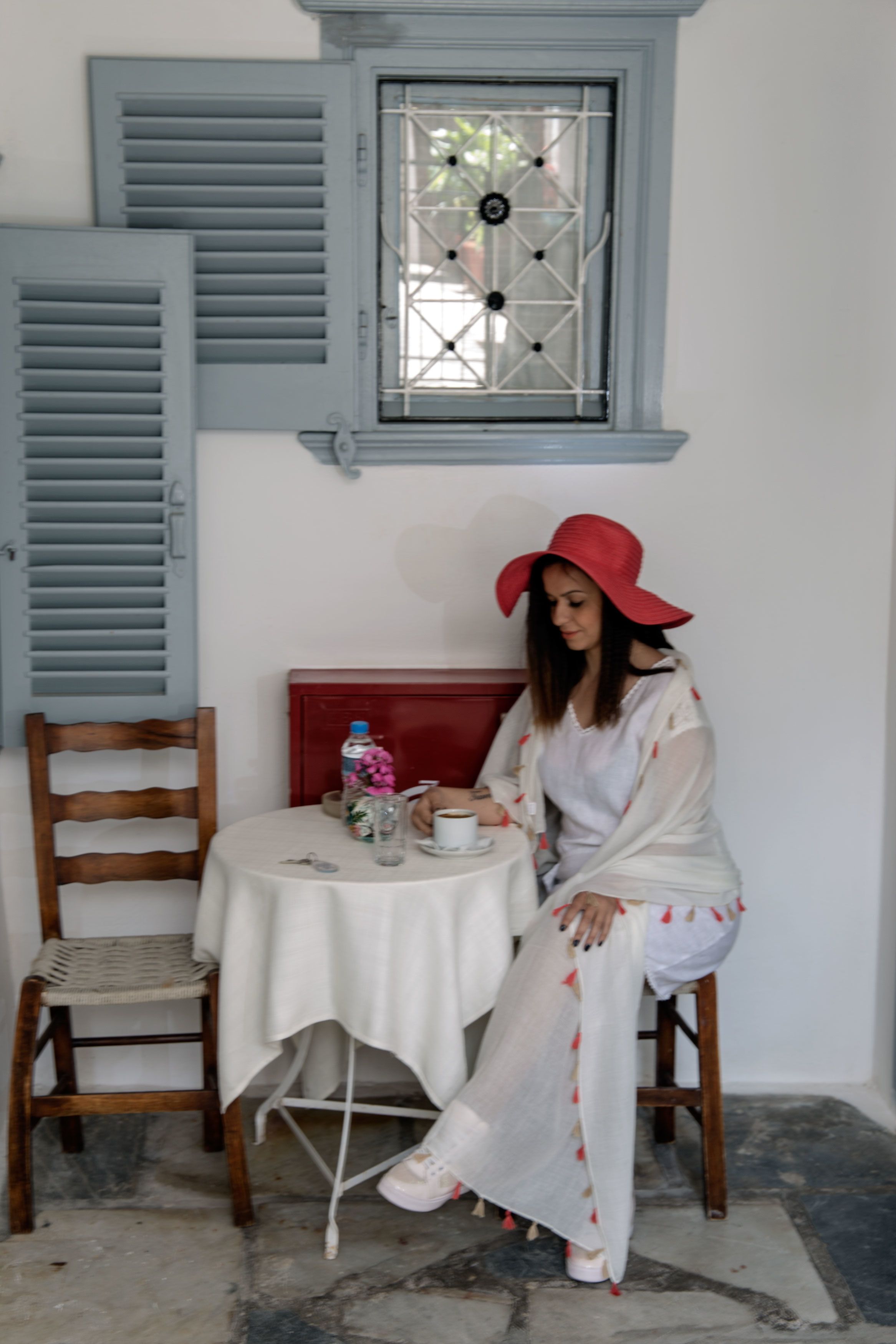 A woman sitting at a table with a white tablecloth, a red chair, and a window with blue shutters.