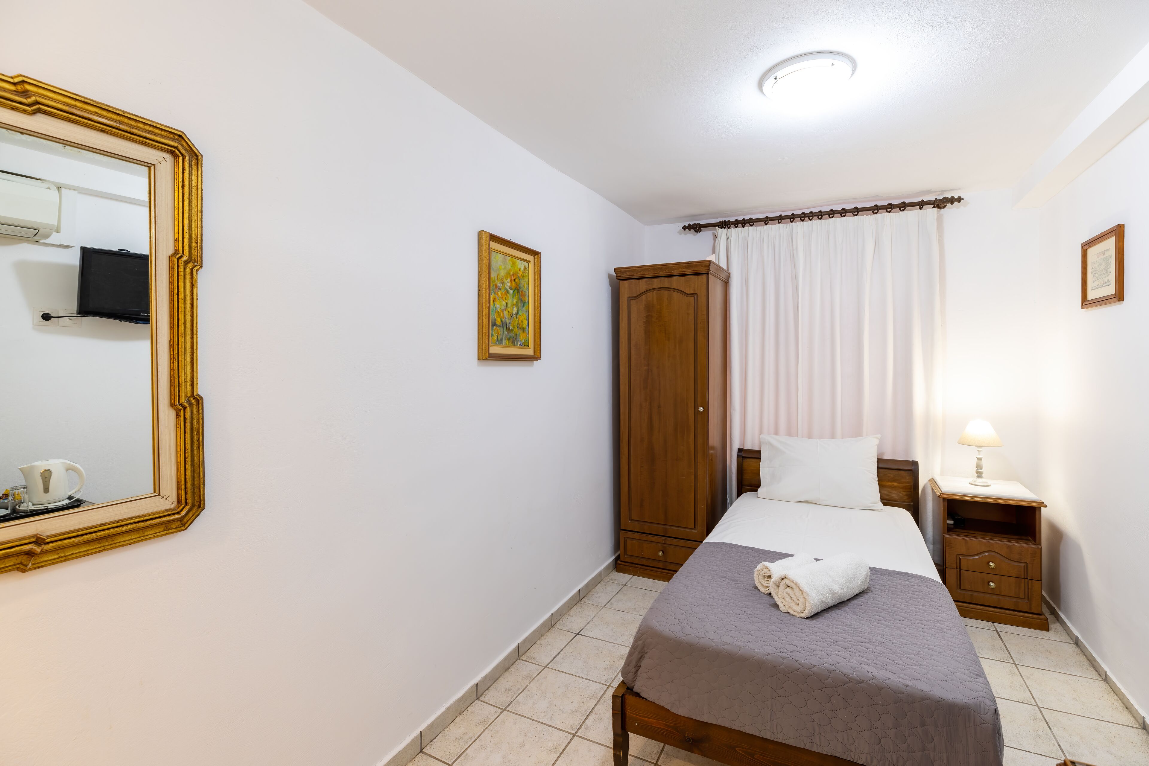 A hotel room with a bed, a wardrobe, a nightstand, a lamp, a mirror, and a painting on the wall.