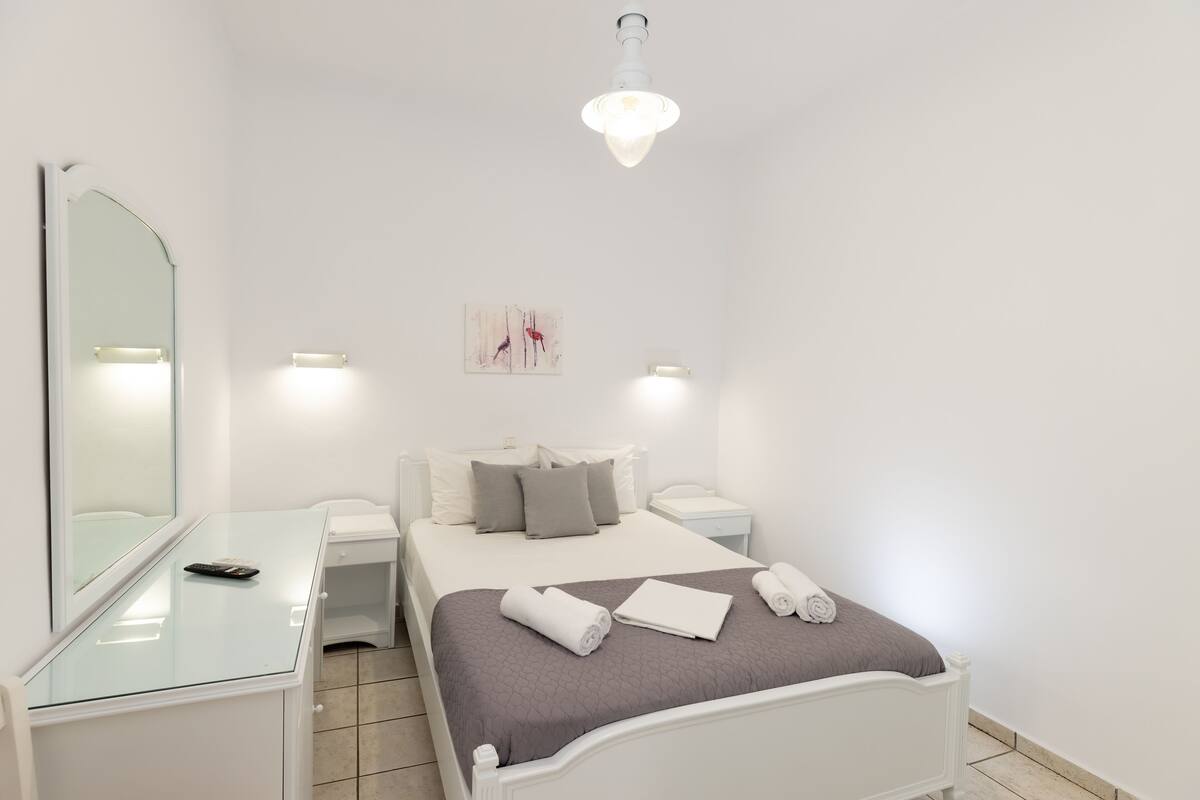 Une chambre moderne avec un bureau à plateau en verre, un lit recouvert d’une couverture grise, deux tables de chevet, un luminaire fixé au mur et une œuvre d’art encadrée.