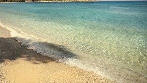 Una playa con agua turquesa cristalina, una orilla arenosa y una costa distante con edificios y árboles.