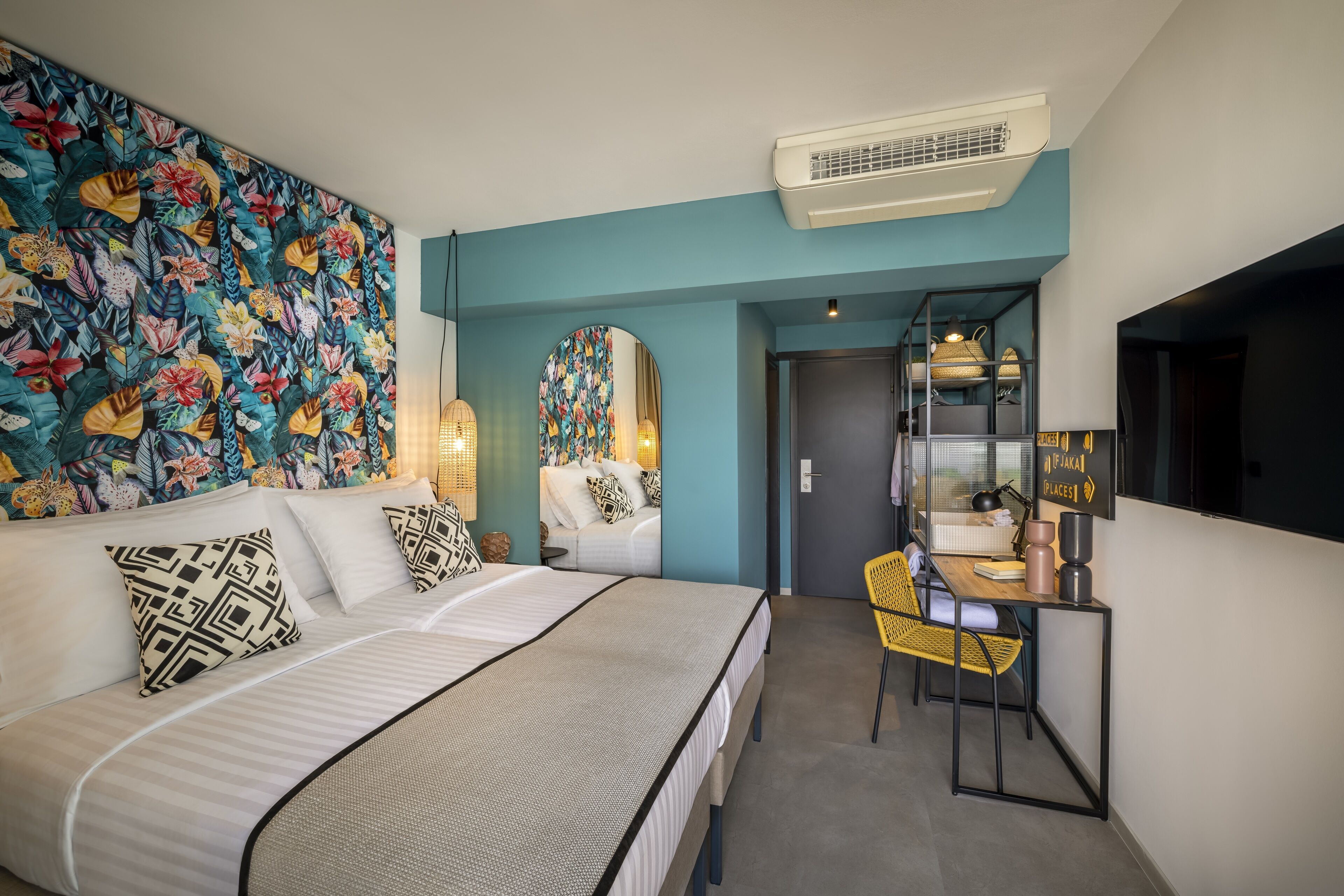 Una habitación de hotel moderna con una cama grande, un escritorio con silla, un tapiz floral llamativo y un espejo.