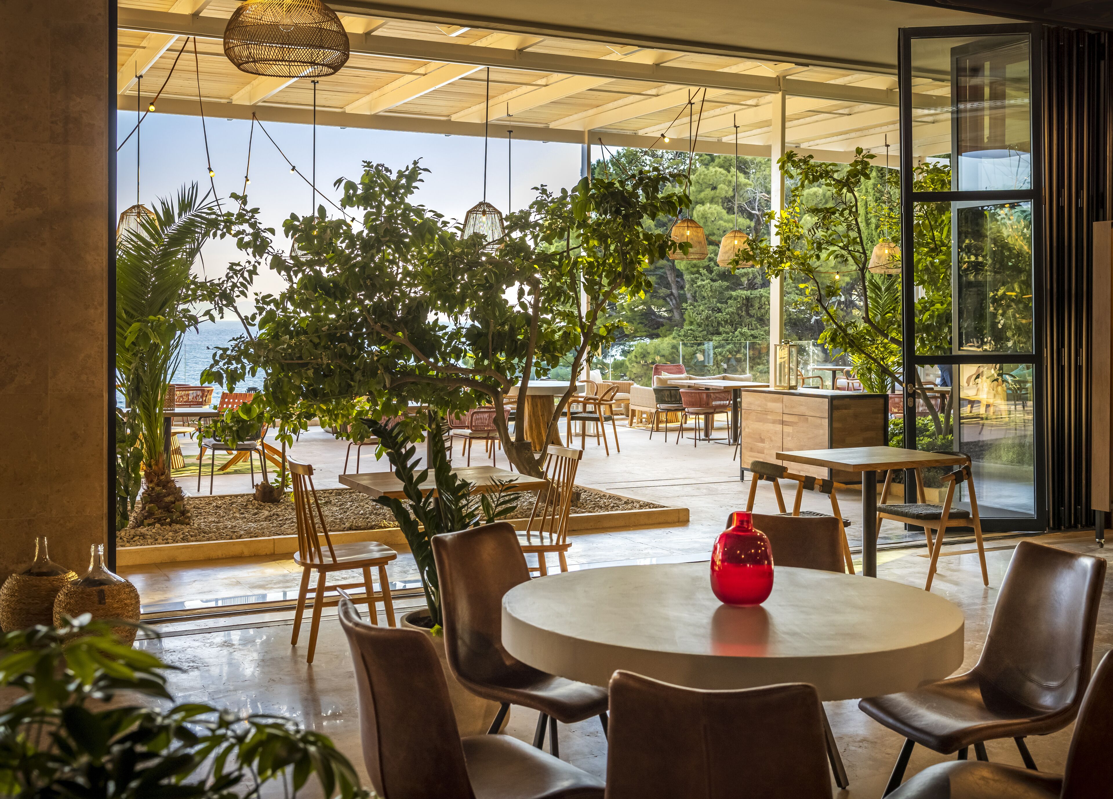 An indoor seating area with round tables and chairs, a large tree, and a view of the sea through large windows.