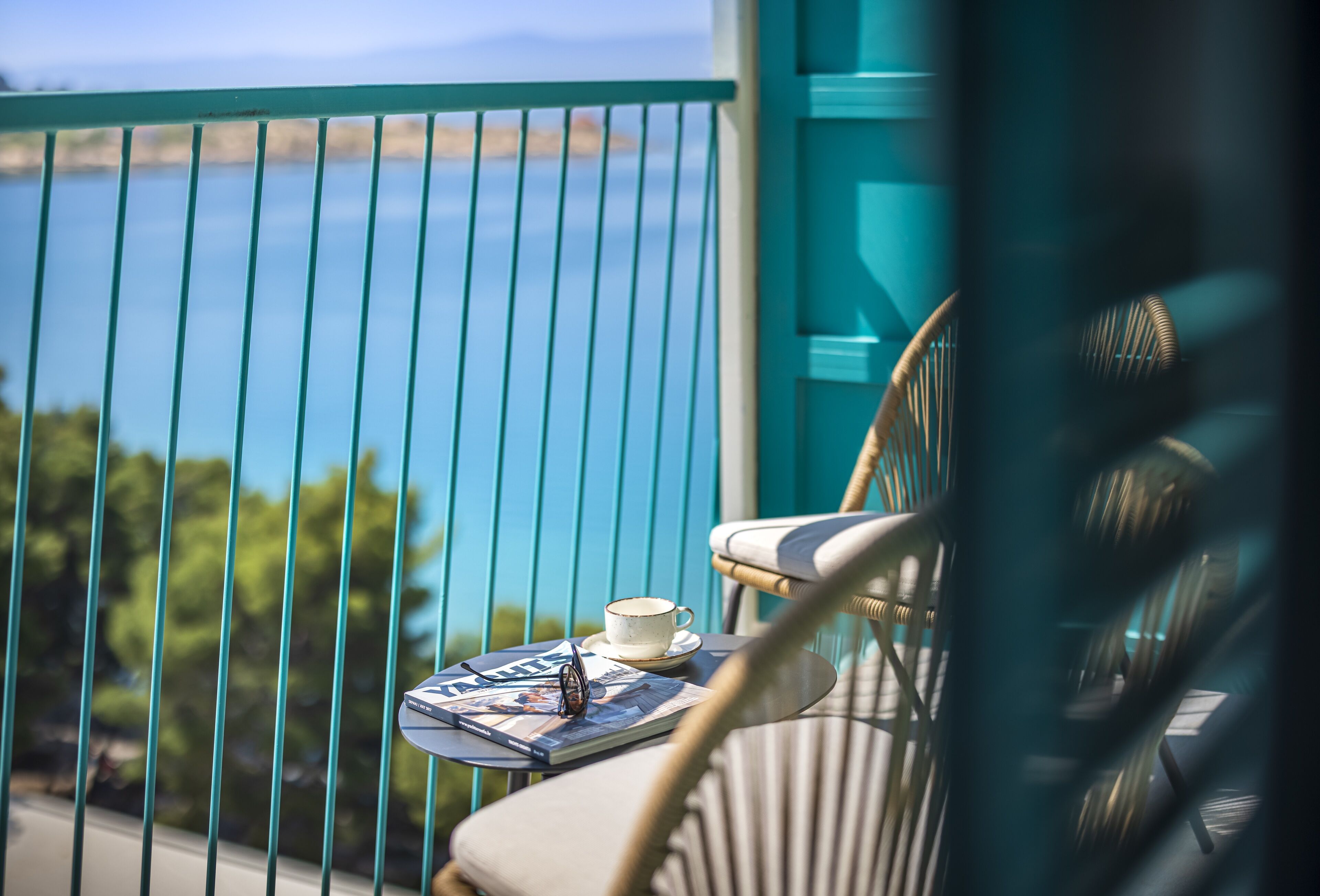 Een balkon met uitzicht op zee, een rieten stoel en een tafel met een kopje en een tijdschrift.