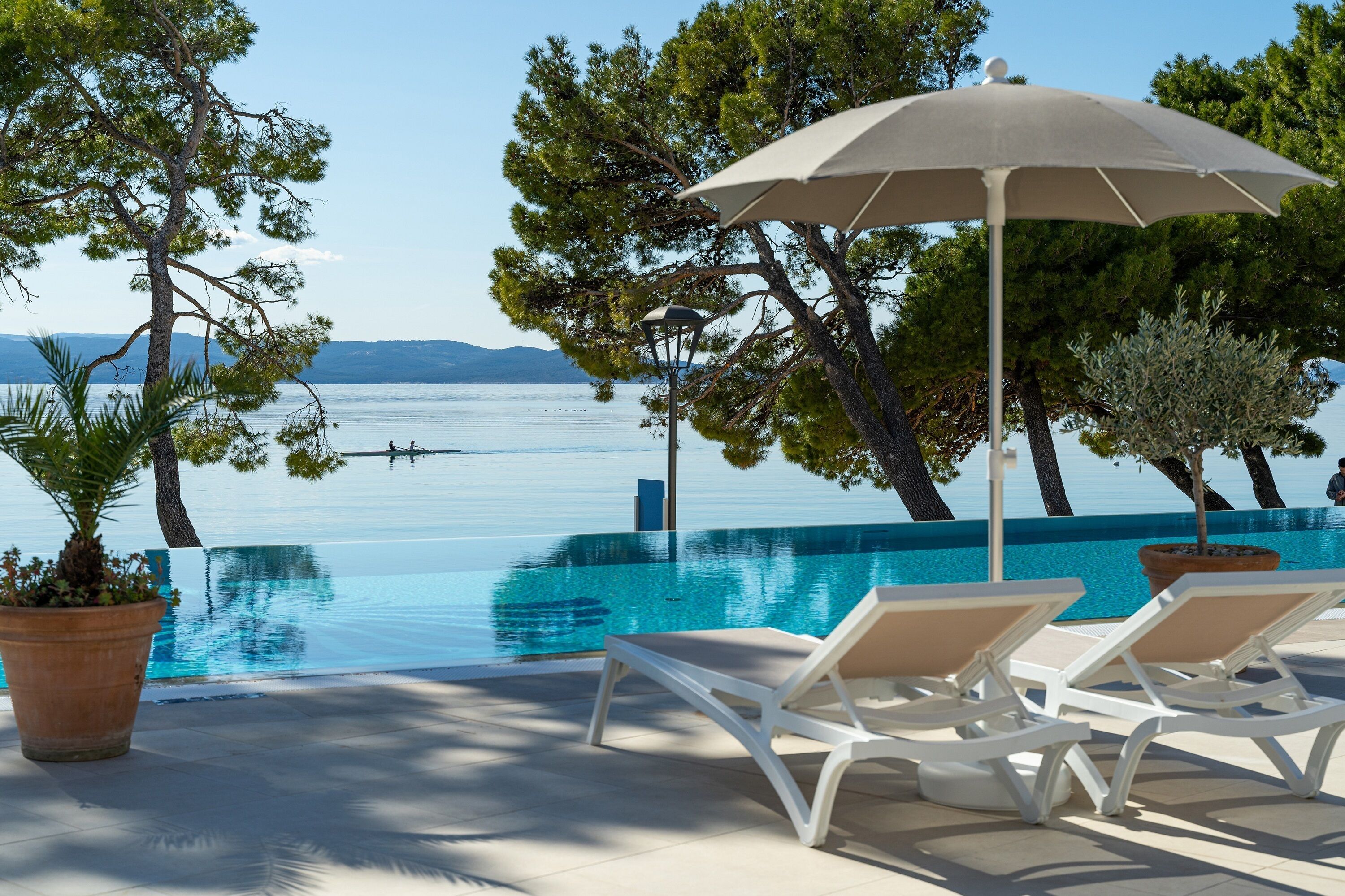 Un espace aménagé au bord de la piscine, avec des chaises longues et un grand parasol, offrant une vue sur une étendue d’eau d’un bleu limpide, avec une terre ferme visible au loin.