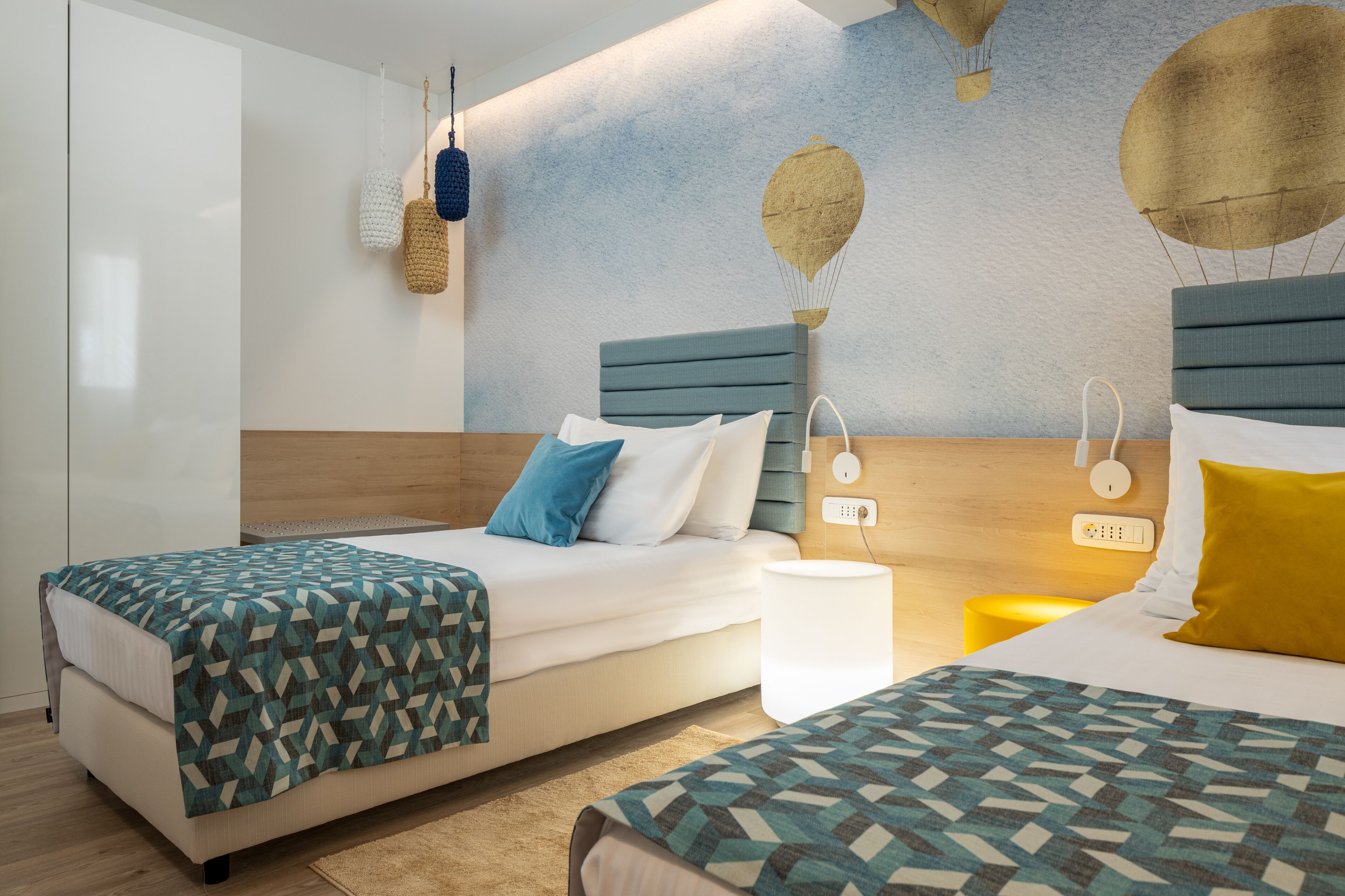 Quarto de hotel moderno com duas camas, cabeceira de madeira, mural na parede com balões de ar quente e mesa de cabeceira com abajur.