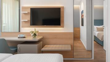 Een moderne hotelkamer met een flatscreen televisie aan de muur, een bureau met stoel, een bed met blauw hoofdeinde en een kleine zithoek met een bankje.