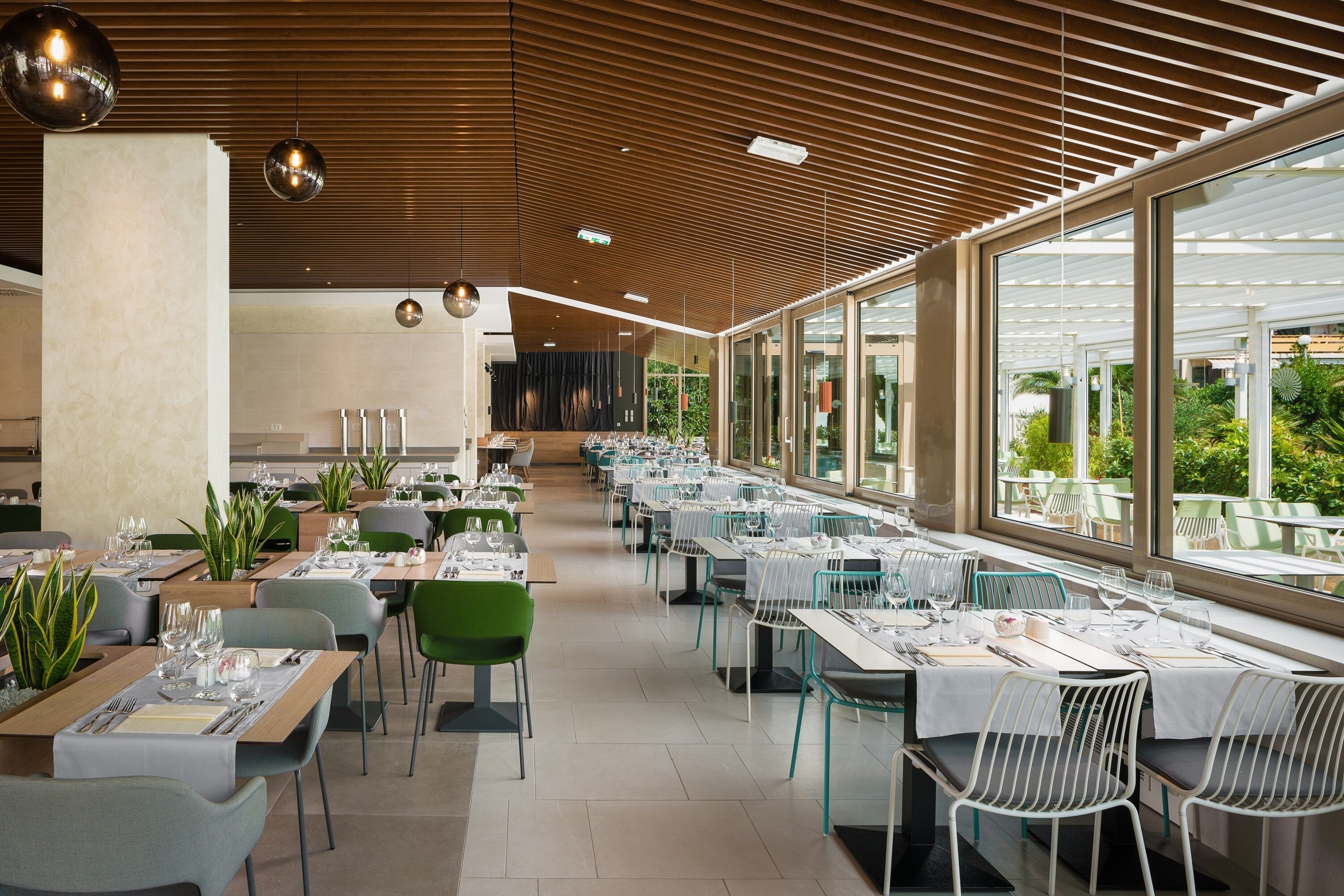 A modern dining area with long tables, green chairs, and large windows offering a view of the outdoors.