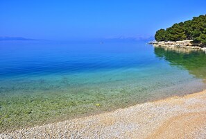 A clear view of a pebble beach leading into calm waters with a distant boat and a forested hill. - Valamar Meteor Hotel (Makarska)
