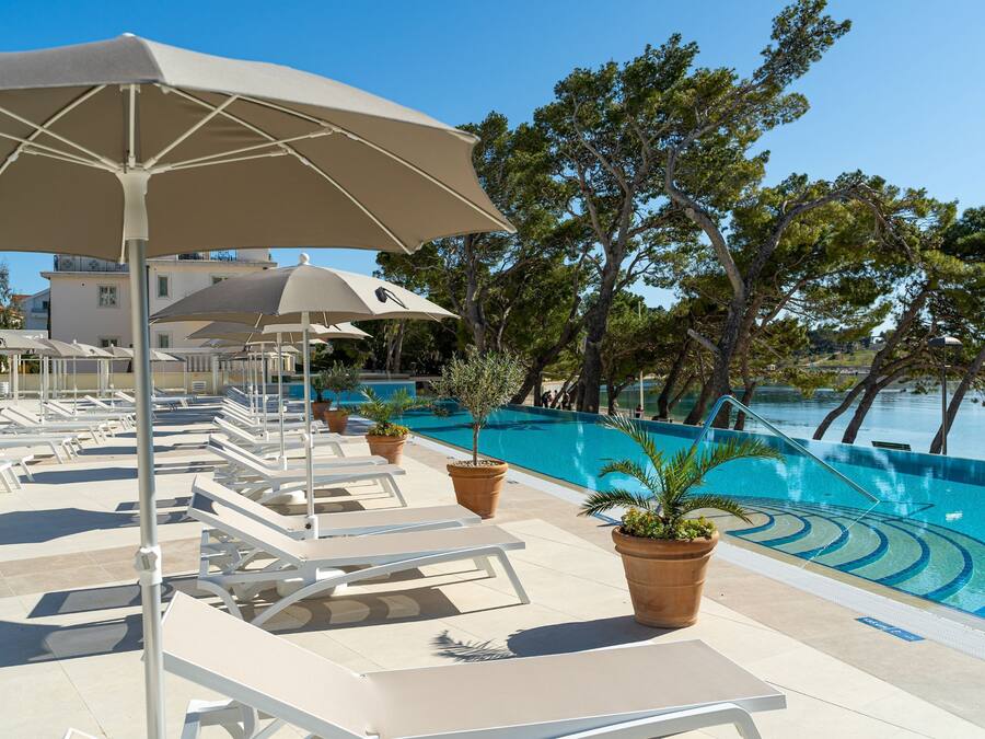 Et poolområde med liggestole og parasoller, omgivet af træer med udsigt over en klar, blå pool.