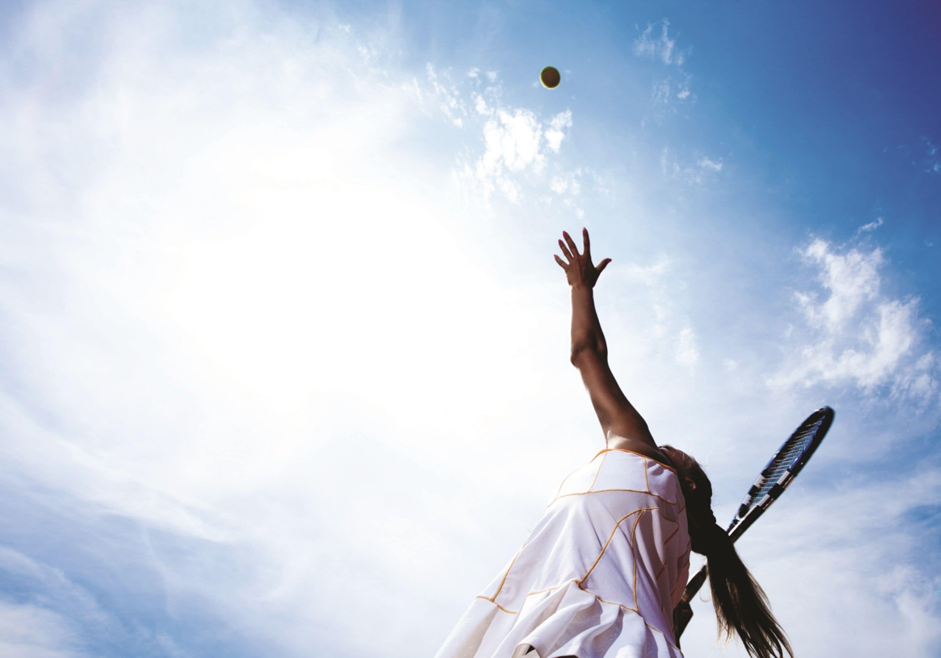Une personne joue au tennis avec une raquette, et tend le bras pour frapper une balle sous un ciel dégagé.