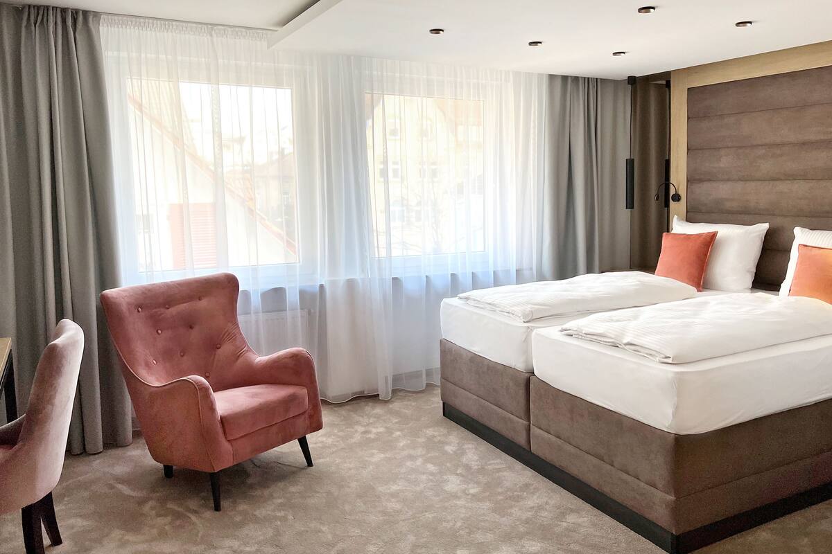 Habitación de hotel con una cama grande, una silla y vistas al exterior a través de cortinas translúcidas.