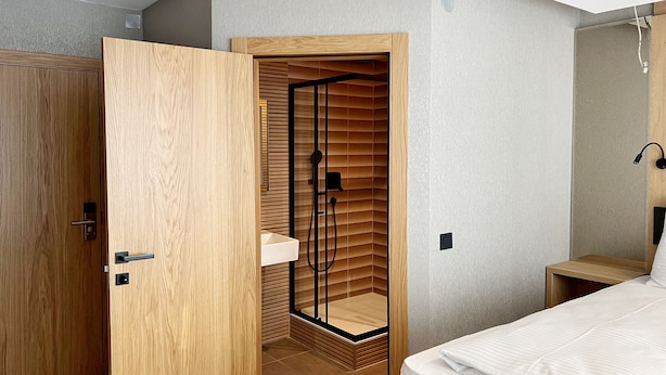 A modern bedroom with a wooden wardrobe, a built-in shower with a glass door, and a bedside table with a lamp. - DORMERO Hotel Reutlingen (Reutlingen)