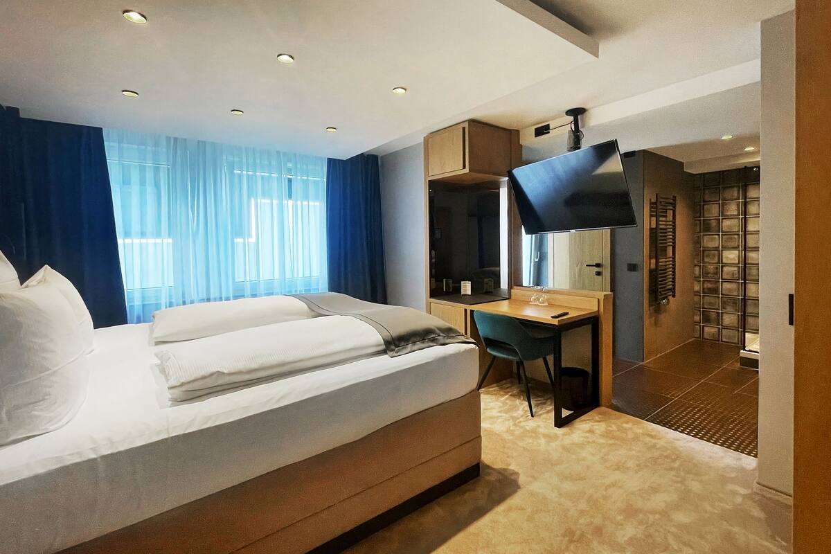 Habitación de hotel con cama, escritorio, silla, televisor y baño con cabina de ducha.