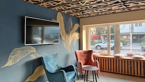 A modern waiting area with a blue wall, a mounted TV, and a window with a view of the street. - DORMERO Hotel Reutlingen (Reutlingen)
