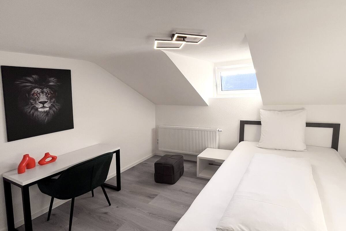 Un dormitorio moderno con una cama blanca, un escritorio, una silla, un radiador y una lámpara en el techo.
