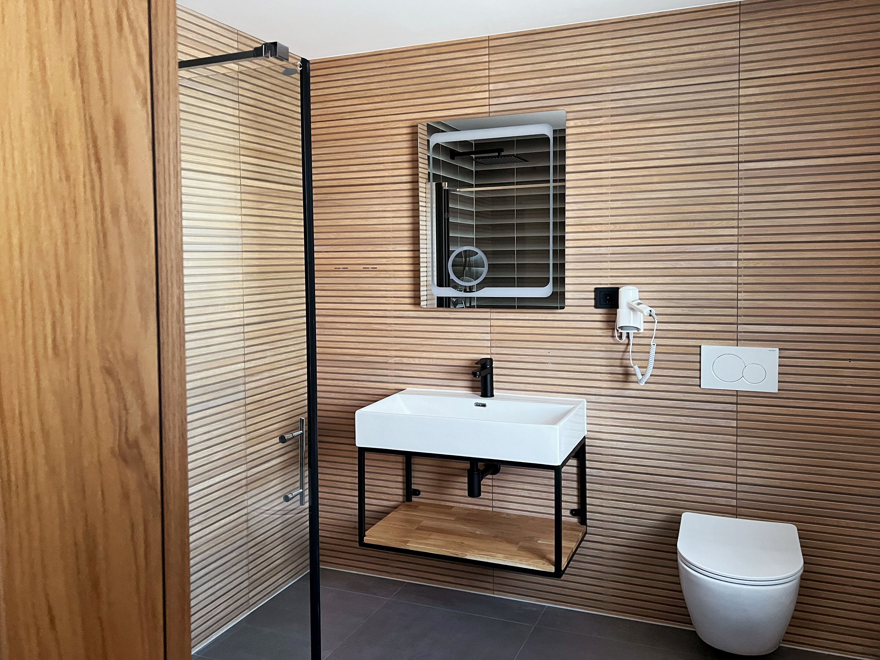 Un bany modern amb paret de panells de fusta, un lavabo blanc, un mirall, una dutxa i un vàter.