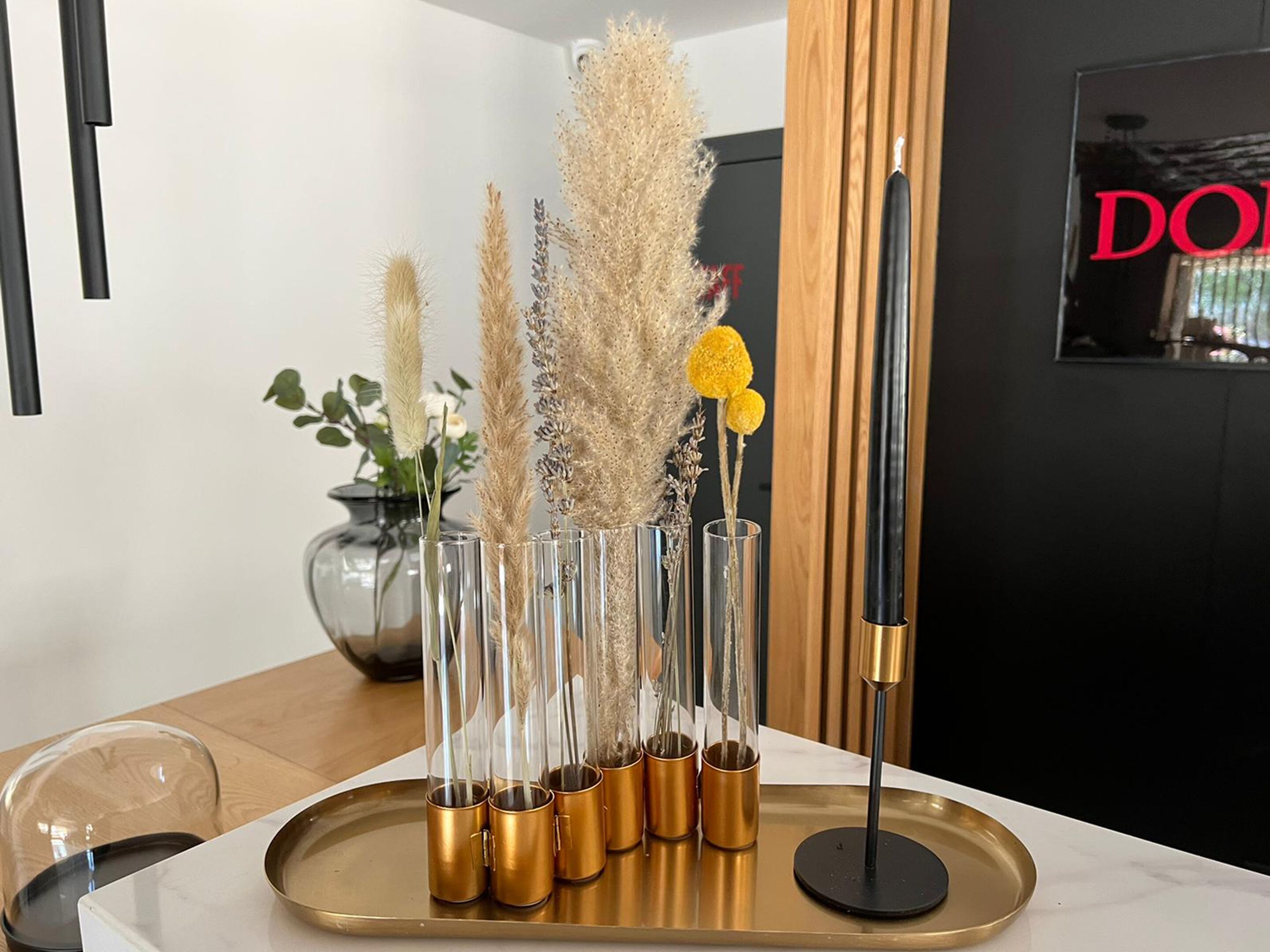 Un espace de salle à manger moderne, agrémenté d’un vase en verre disposé sur un plateau, d’un grand lampadaire noir et doré, et d’un panneau en bois en arrière-plan.