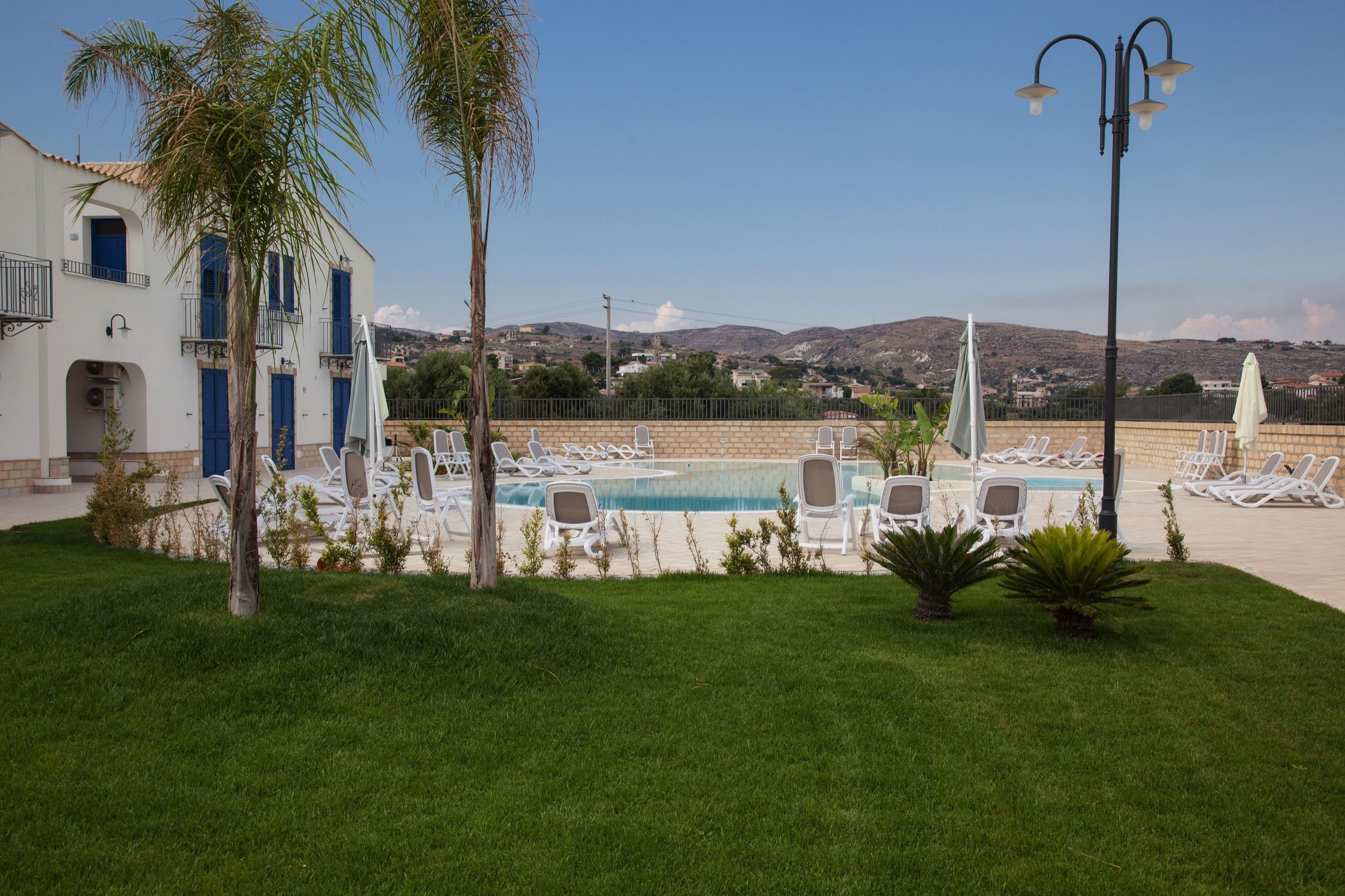 Zona de piscina con sillones de descanso, una palmera y un edificio con puertas y ventanas azules.